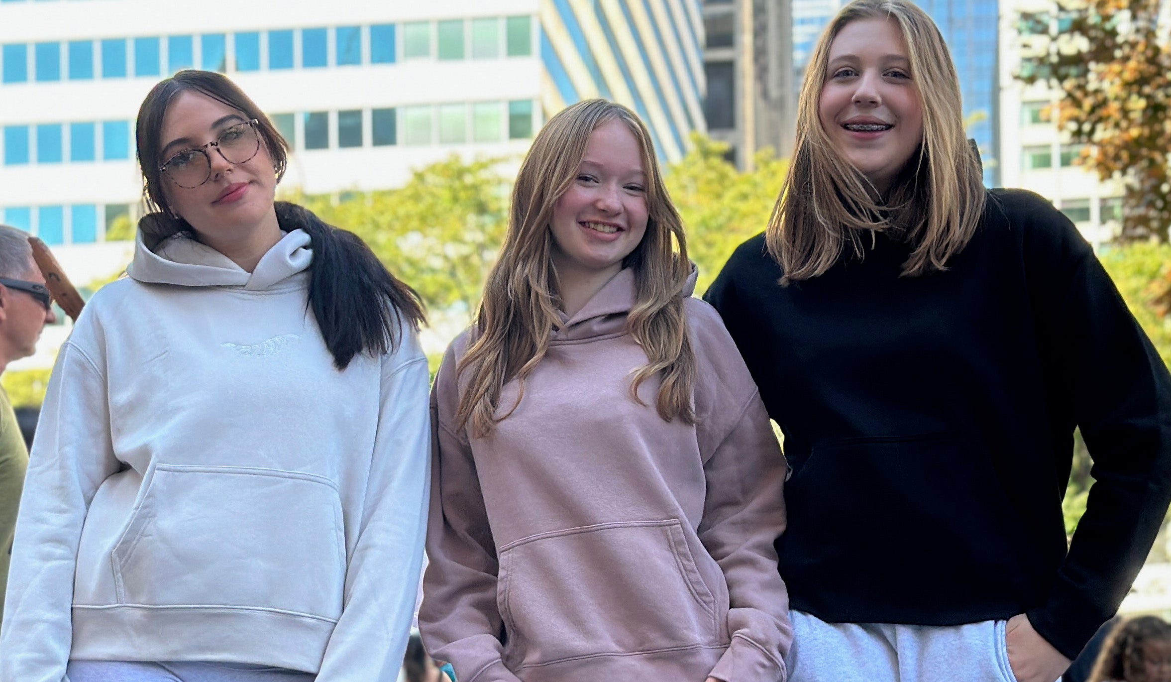 Bone coloured, hazy pink and black hoodies worn by 3 women in front of a city scape. Normalize Neurodivergence.