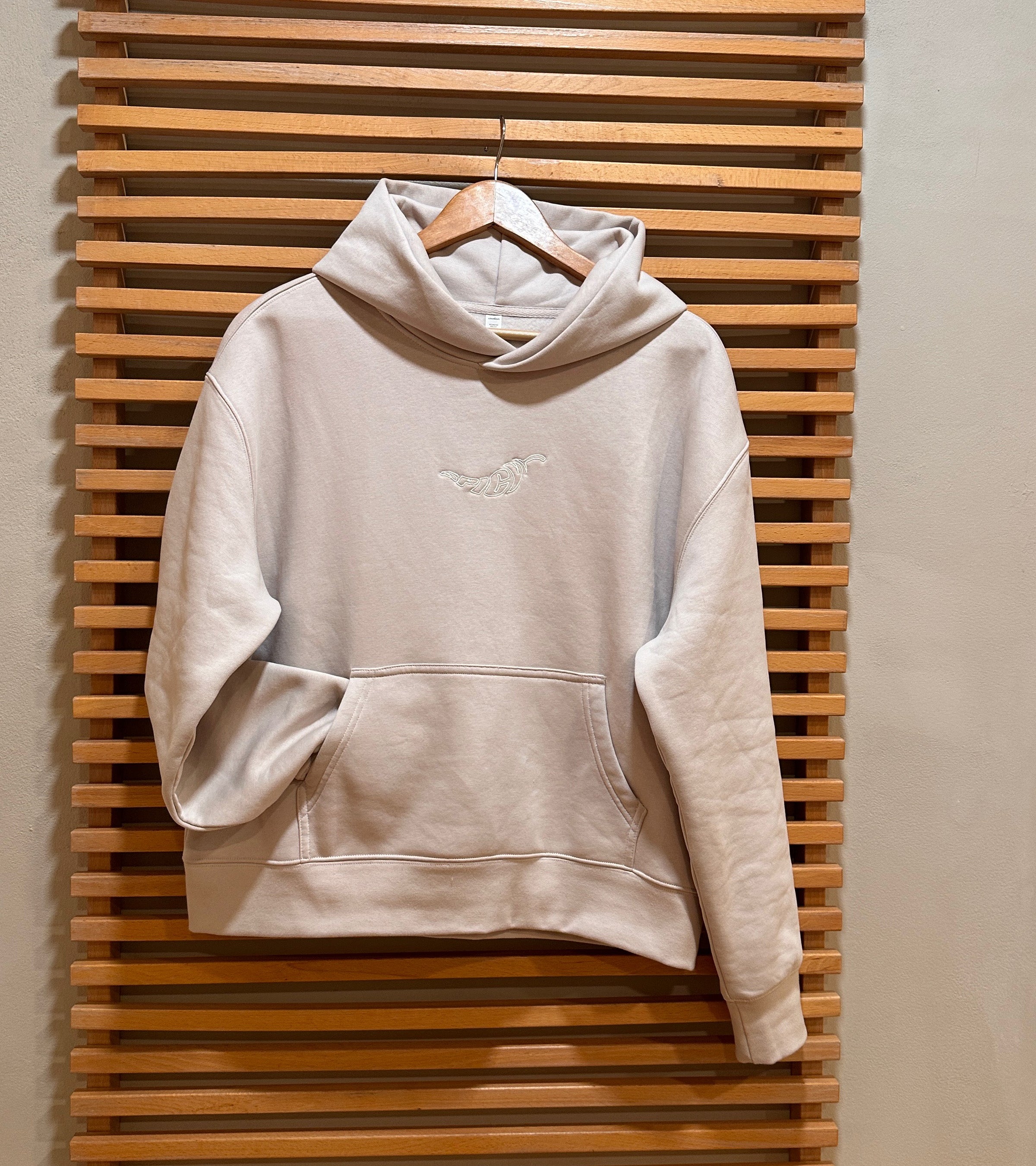 Bone colored women's hoodie on hanger against a slatted wall. Embroidered spicy logo on hoodie center front. Normalize Neurodivergence Movement